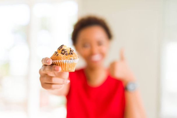 Young african american woman eating chocolate chips muffins happy with big smile doing ok sign, thumb up with fingers, excellent sign - Fotografie, Obrázek