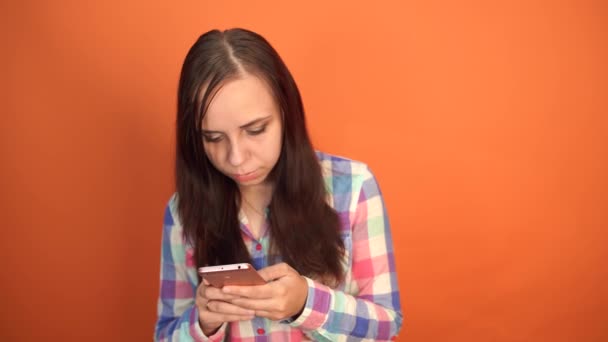 Young woman holding mobile phone in hand on orange background. Female typing a message on a smartphone - Materiał filmowy, wideo