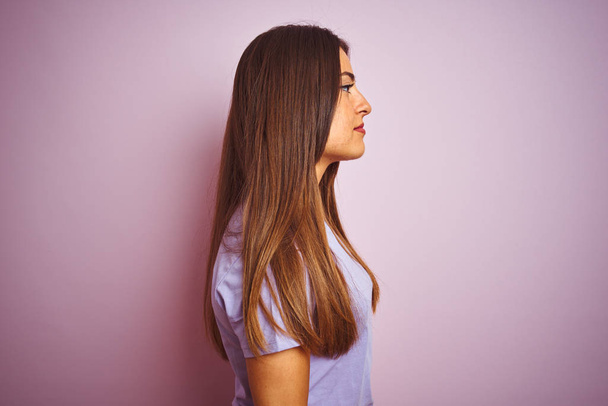 Mladá krásná žena, která měla na sobě neformální tričko stojící nad osamělým růžovým pozadím, která se dívá na stranu, odpočívá v profilu s přirozenou tváří s důvěřným úsměvem. - Fotografie, Obrázek