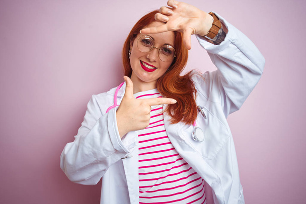 Νεαρή κοκκινομάλλα γιατρός γυναίκα φορώντας γυαλιά πάνω από ροζ απομονωμένο φόντο χαμογελώντας καθιστώντας το πλαίσιο με τα χέρια και τα δάχτυλα με χαρούμενο πρόσωπο. Φιλοσοφία δημιουργικότητας και φωτογραφίας. - Φωτογραφία, εικόνα