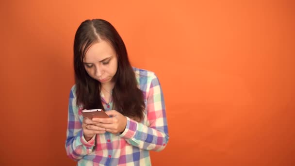 Young woman holding mobile phone in hand on orange background. Female typing a message on a smartphone - Imágenes, Vídeo