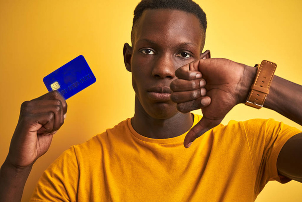Homme client afro-américain tenant la carte de crédit debout sur fond jaune isolé avec le visage en colère, signe négatif montrant aversion avec les pouces vers le bas, concept de rejet
 - Photo, image