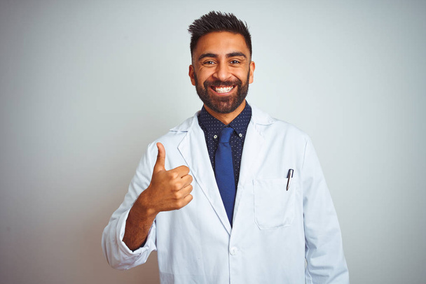 Young indian doctor man standing over isolated white background doing happy thumbs up gesture with hand. Approving expression looking at the camera with showing success. - Photo, Image