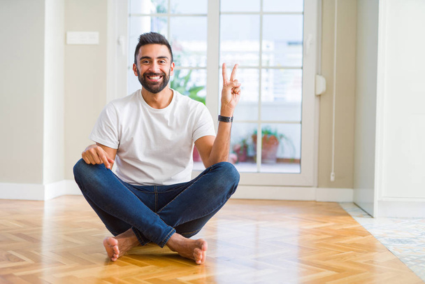 Handsome hispanic man wearing casual t-shirt sitting on the floor at home smiling with happy face winking at the camera doing victory sign. Number two. - Photo, Image