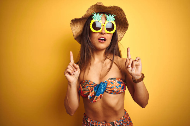 Woman on vacation wearing bikini and pineapple sunglasses over isolated yellow background amazed and surprised looking up and pointing with fingers and raised arms. - Valokuva, kuva