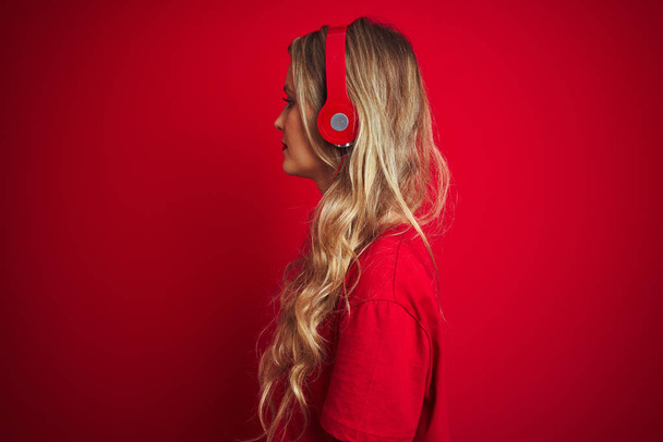 横を見て赤い孤立した背景の上にヘッドフォンを着用した若い美しい女性は、自信のある笑顔で自然な顔でプロフィールポーズをリラックス. - 写真・画像