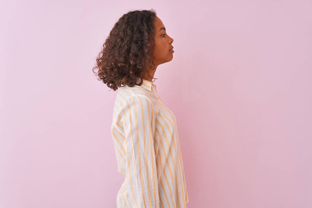Jovem brasileira vestindo camisa listrada em pé sobre fundo rosa isolado olhando para o lado, relaxar pose perfil com rosto natural com sorriso confiante
. - Foto, Imagem
