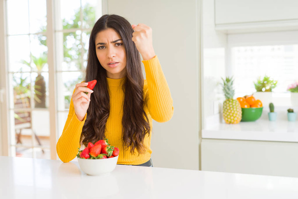 Mujer joven comiendo fresa roja fresca como bocadillo saludable molesto y frustrado gritando con ira, loco y gritando con la mano levantada, concepto de ira
 - Foto, imagen
