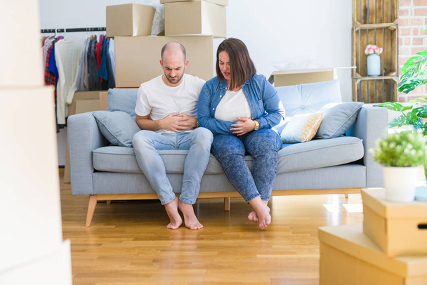 Young couple sitting on the sofa arround cardboard boxes moving to a new house with hand on stomach because indigestion, painful illness feeling unwell. Ache concept. - Photo, Image