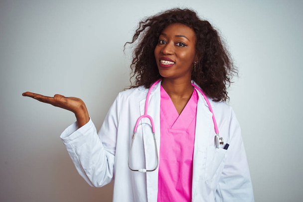 Africká americká doktorka, Žena s růžovým stetoskem nad osamělým bílým pozadím, usměvavá a s dlaní, ukazující na kameru. - Fotografie, Obrázek