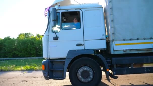 Truck met vracht trailer rijden op de snelweg en het vervoeren van goederen. Witte vrachtwagen rijden naar bestemming door landelijke weg op zonnige dag. Felle zon schijnt op de achtergrond. Zijaanzicht Slow Motion - Video