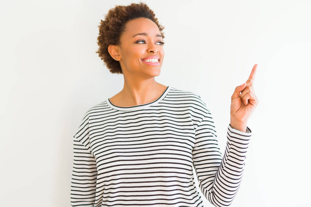 Jovem mulher americana africana bonita vestindo listras suéter sobre fundo branco com um grande sorriso no rosto, apontando com a mão e o dedo para o lado olhando para a câmera
. - Foto, Imagem