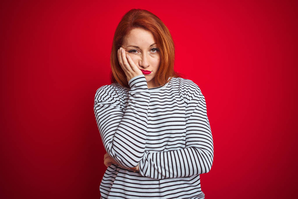 赤い孤立した背景の上に立つストラップネイビーシャツを着た若い赤毛の女性は、腕を組んだうつ病の問題に疲れて退屈しているように見える. - 写真・画像