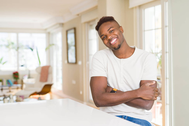 gutaussehender afrikanisch-amerikanischer Mann auf weißem Tisch zu Hause, fröhliches Lächeln mit verschränkten Armen, die in die Kamera schauen. positive Person. - Foto, Bild