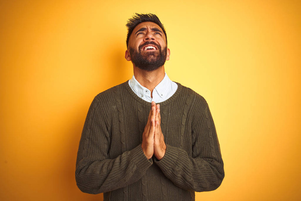 Young indian man wearing green sweater and shirt standing over isolated yellow background begging and praying with hands together with hope expression on face very emotional and worried. Asking for forgiveness. Religion concept. - Photo, Image