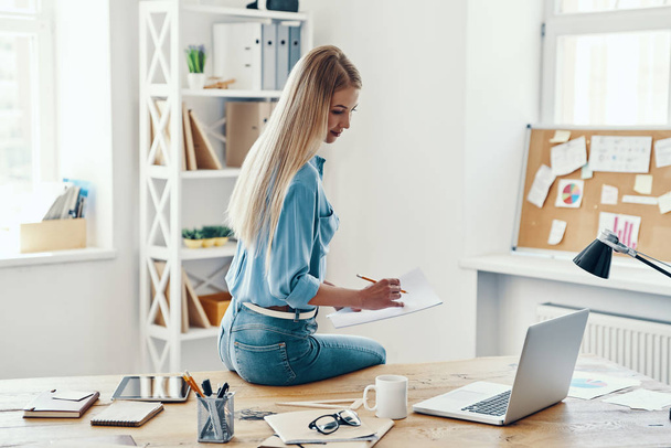 Beautiful young woman in smart casual wear writing something down and using laptop while sitting in the office - Foto, imagen