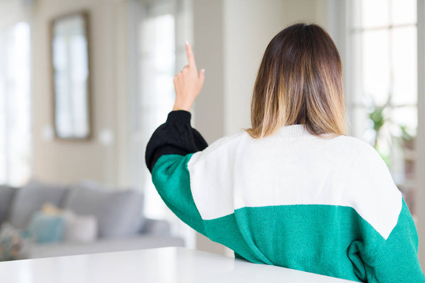 Young beautiful woman wearing winter sweater at home Posing backwards pointing behind with finger hand - Fotó, kép
