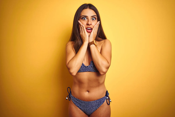 Young beautiful woman on vacation wearing bikini standing over isolated yellow background afraid and shocked, surprise and amazed expression with hands on face - Photo, Image