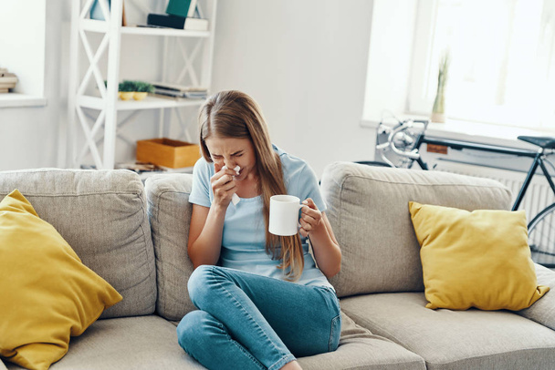 Ill young woman blowing nose and sneezing while sitting on the sofa at home           - Photo, image