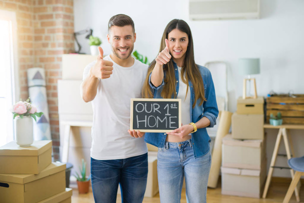 Young beautiful couple holding blackboard with our first home text at new house happy with big smile doing ok sign, thumb up with fingers, excellent sign - Fotó, kép