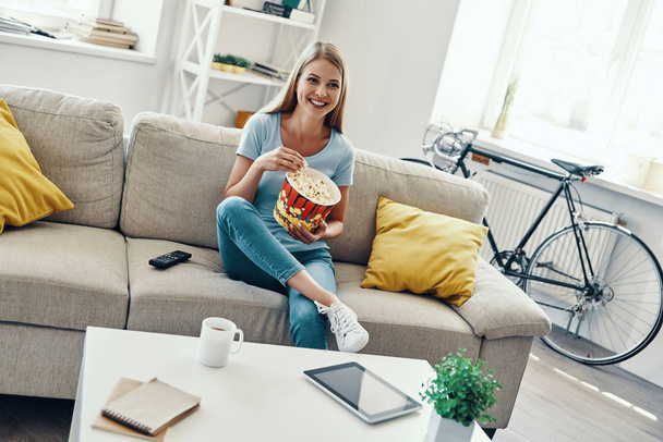 Beautiful young woman eating popcorn and smiling while watching TV on the sofa at home               - Foto, afbeelding