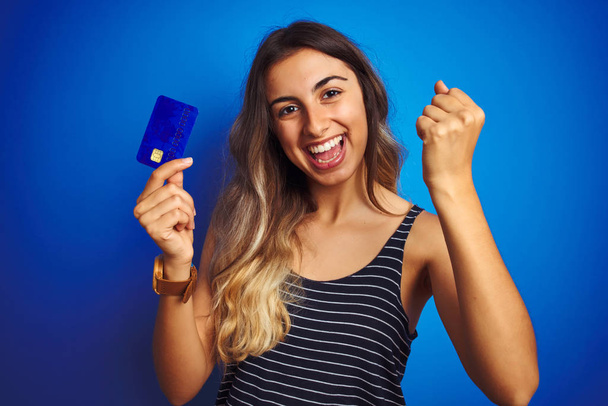 Young beautiful woman holding credit card over blue isolated background screaming proud and celebrating victory and success very excited, cheering emotion - Photo, Image