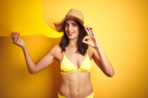 Beautiful woman wearing yellow bikini and holding talking balloon over isolated yellow background doing ok sign with fingers, excellent symbol - Photo, Image