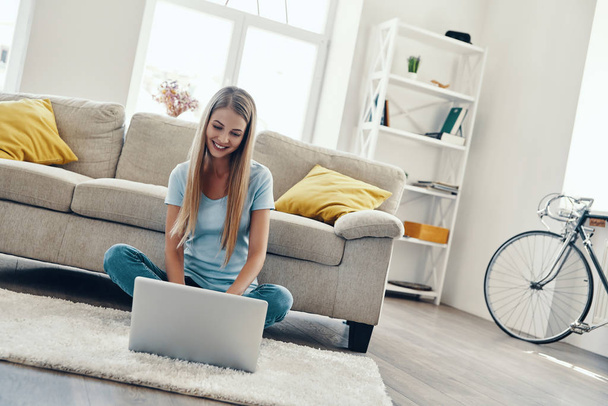Beautiful young woman smiling and using laptop while resting at home        - Foto, Imagen