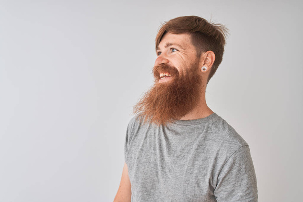 Nuori punapää irlantilainen mies päällään t-paita seisoo yli eristetty harmaa tausta etsii sivuun hymyillen kasvot, luonnollinen ilme. Nauraen itsevarmana
. - Valokuva, kuva