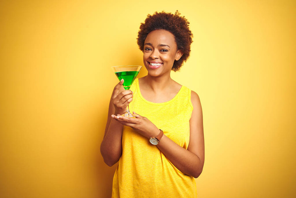 Jeune femme afro-américaine aux cheveux afro buvant un cocktail sur fond jaune isolé avec un visage heureux debout et souriant avec un sourire confiant montrant des dents
 - Photo, image