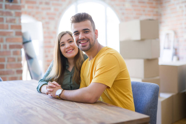Young beautiful couple sitting on the table at home, hugging in love very happy for moving to new home with cardboard boxes behind them - Photo, Image