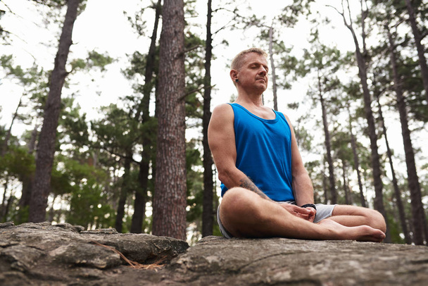 Man sitting with his legs crossed and eyes closed on a rock and meditating while practicing yoga alone in a pine forest - Фото, изображение