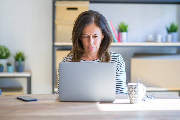 Middle age senior woman sitting at the table at home working using computer laptop skeptic and nervous, frowning upset because of problem. Negative person. - 写真・画像