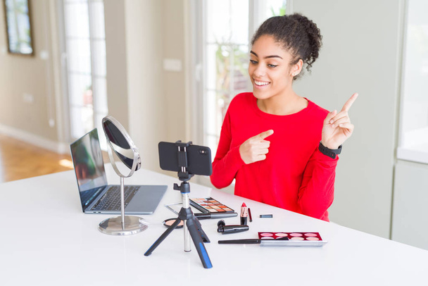 junge afrikanisch-amerikanische Influencerin nimmt Make-up-Anleitung lächelnd auf und blickt in die Kamera, die mit zwei Händen und Fingern zur Seite zeigt. - Foto, Bild