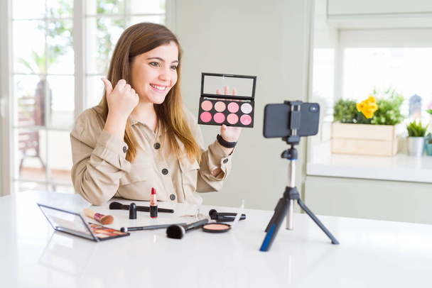 Mooie jonge influencer vrouw opname make-up video tutorial aanwijzen en tonen met duim aan de zijkant met vrolijk gezicht glimlachend - Foto, afbeelding