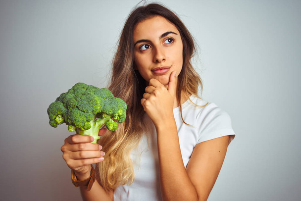 Młoda piękna kobieta jedzenia brokułów nad szarym białym tle poważne myślenie o pytanie, bardzo pomylony pomysł - Zdjęcie, obraz