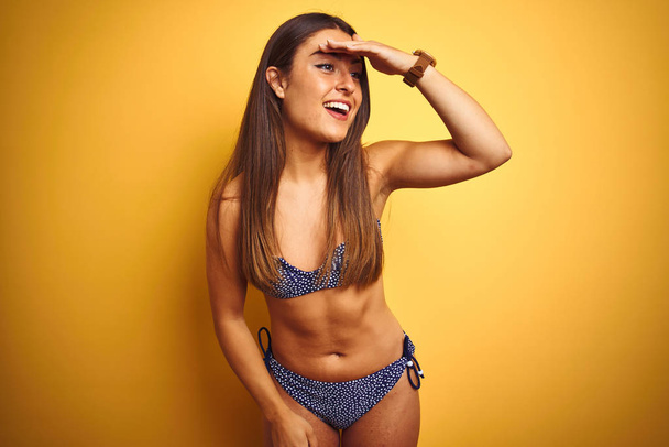 Young beautiful woman on vacation wearing bikini standing over isolated yellow background very happy and smiling looking far away with hand over head. Searching concept. - Photo, Image