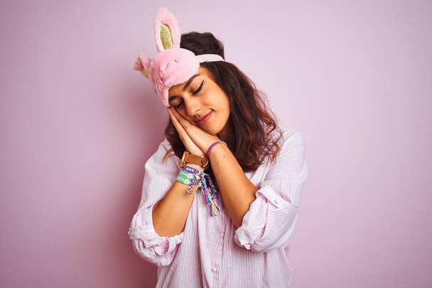 Jovem mulher bonita vestindo pijama e máscara de sono sobre fundo rosa isolado dormindo cansado sonhando e posando com as mãos juntas enquanto sorri com os olhos fechados
. - Foto, Imagem