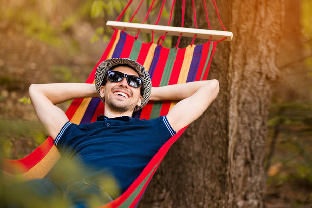 ハンモックで休みながら、森の自然に囲まれた腕を上げる帽子とサングラスをかけた気楽で自由な男 - 写真・画像