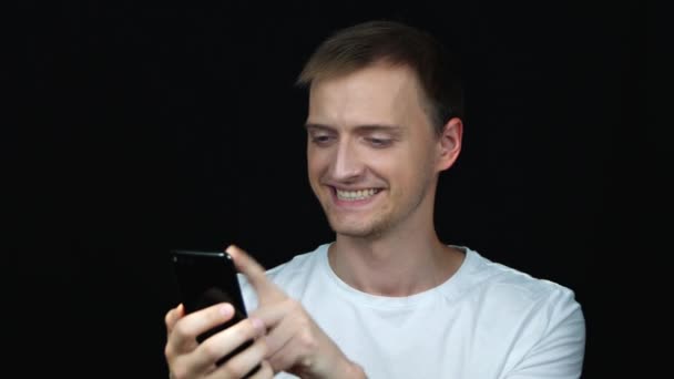 Lachen en lachen jonge blanke man in wit t-shirt met behulp van smartphone over zwarte achtergrond. Close-up staand.  - Video