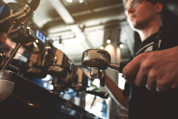 barista man in glasses holding coffee holder with ground coffee in one hand standing near professional coffee machine preparing coffee drink in cafe - Photo, image