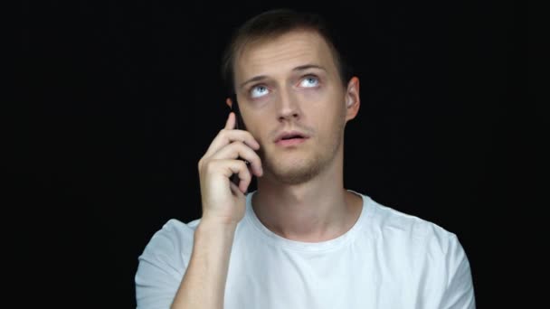 junger Mann irritiert und müde, während er auf einem Mobiltelefon vor schwarzem Hintergrund spricht. Porträt.  - Filmmaterial, Video