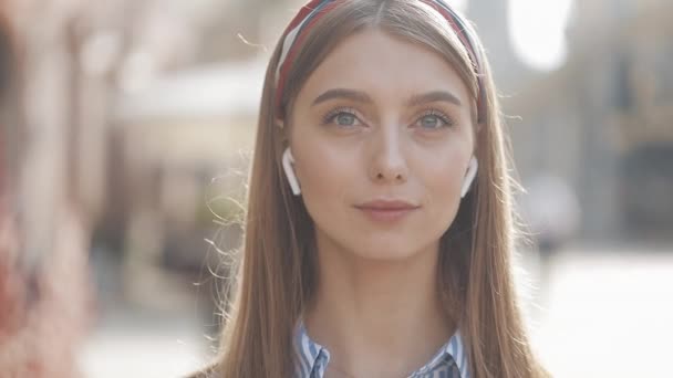 Portrait of a beautiful young woman with wireless earphones looking with into the camera and smiling standing on the old street background. Girl wearing in striped shirt dress with headband. - Filmagem, Vídeo