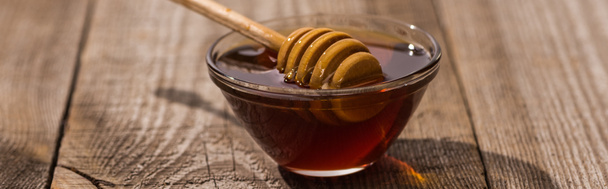panoramic shot of glass bowl with honey and honey dipper on wooden table in sunlight - Foto, Imagen