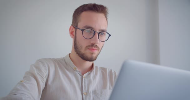 Closeup portrait of young handsome caucasian businessman in the eyeglasses typing on the laptop looking at camera indoors in the white apartment - 映像、動画