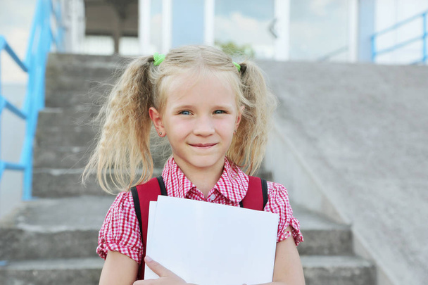 Возвращайся в школу. Beautiful blond schoolgirl with red backpack and books outside the primary school, education concept
. - Фото, изображение