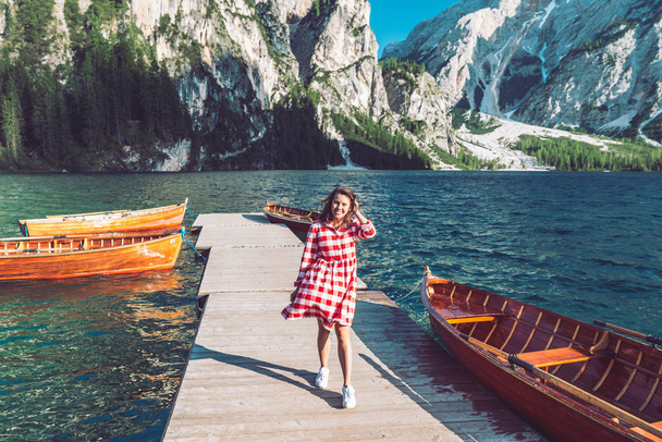 woman walking by mountain lake pier with wooden boats in red dress - Photo, Image