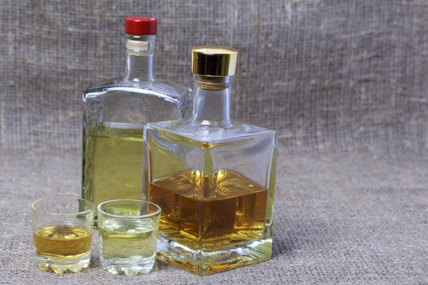 Bottled bottles of strong alcohol. From transparent glass, a square form. Near the stack with a drink poured. Against the backdrop of coarse linen fabric. - Foto, Bild