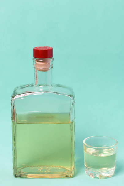 A bottle of strong alcohol. From transparent glass, a square form with a red stopper. Near a stack with a drink poured. Against the background of mint color. - Foto, immagini