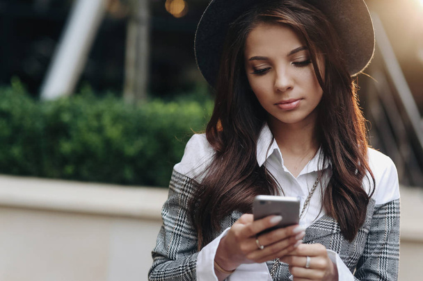 Close up portrait of good-looking woman with modern black hat using her smartphone playing games on mobile phone - Photo, Image